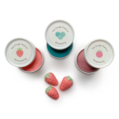Three tubs of dough (strawberry, blueberry, raspberry) on white background with sculpted strawberries out of dough