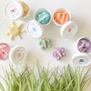 5 tubs of colourful dough on white table with grass decoration at the bottom, sun and butterflies sculpted from dough sit on the table