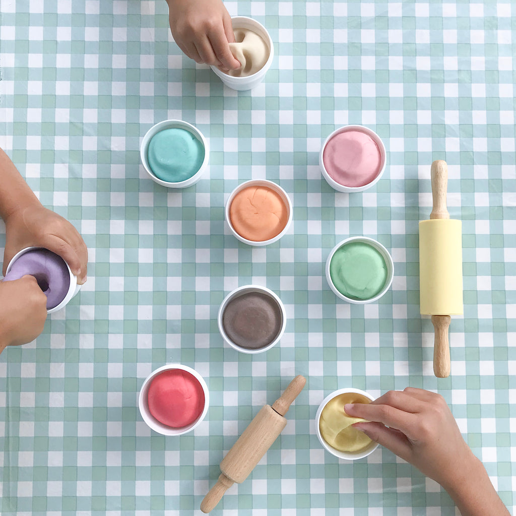 checkerboard table with rolling pins and multiple pots of colourful dough, children's hands squishing the dough in the pots