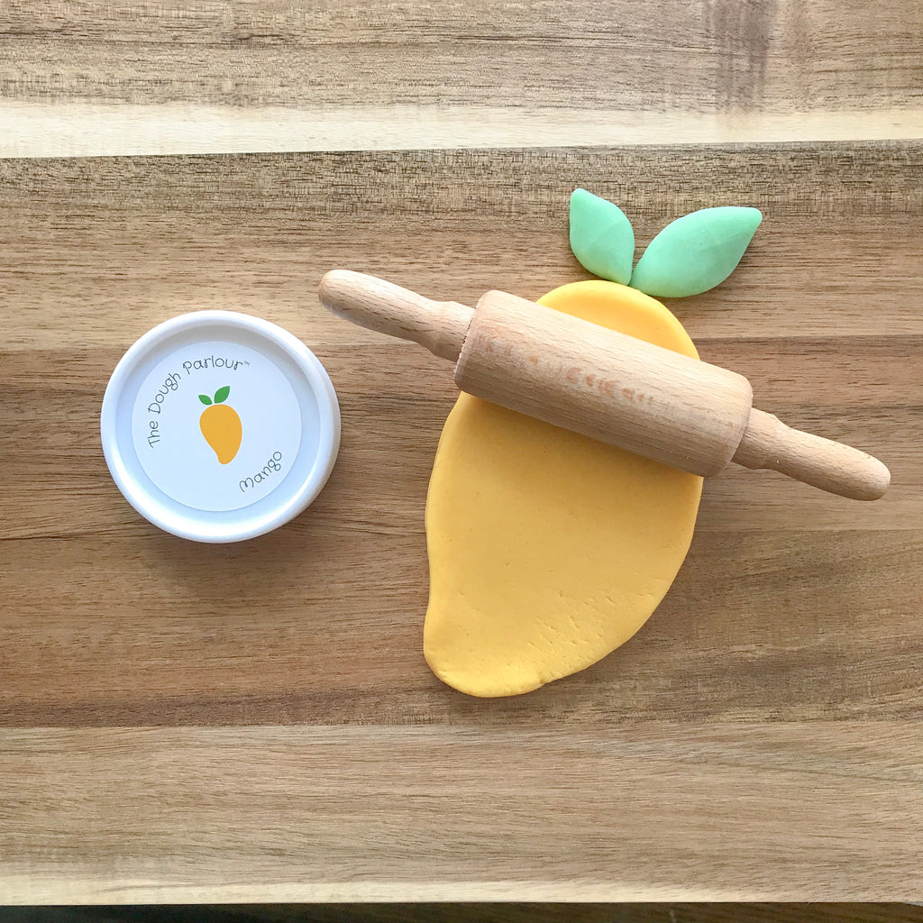 The Dough Parlour mango scented orange dough rolled out with a wooden rolling pin in the shape of a mango beside a tub