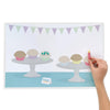 Birthday mat printing shows green and purple bunting with two cake plates holding three cupcakes on each, sitting on a blue table. A child's hand holds a modelling dough candle they placed over one cupcake