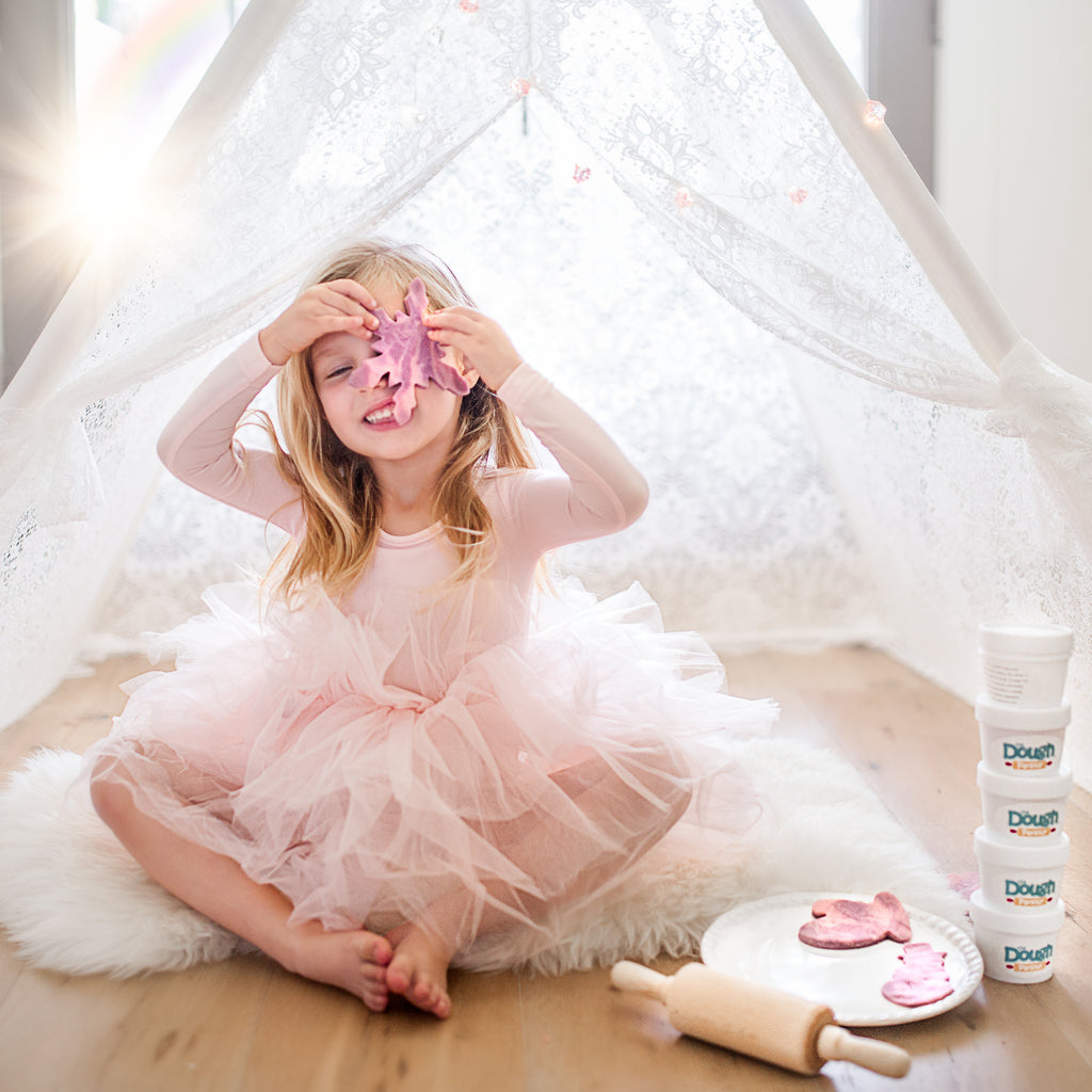 Little girl in pink tutu sitting on faux fur rug under white tent, playing with dough cut outs with a stack of five dough tubs beside her