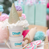 Two tubs of dough stacked with a glitter party hat on top, background shows candy and party decorations