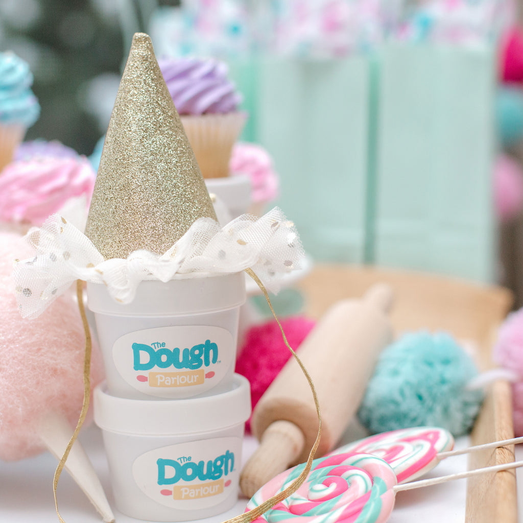 Two tubs of dough stacked with a glitter party hat on top, background shows candy and party decorations