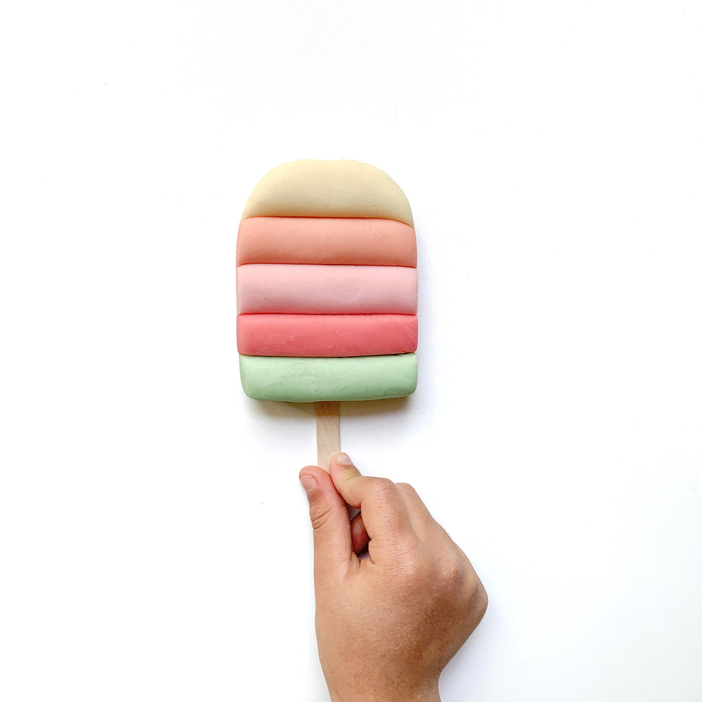 Child's hand holding wooden stick with dough sculpted into ice cream treat with five different stripes of coloured dough