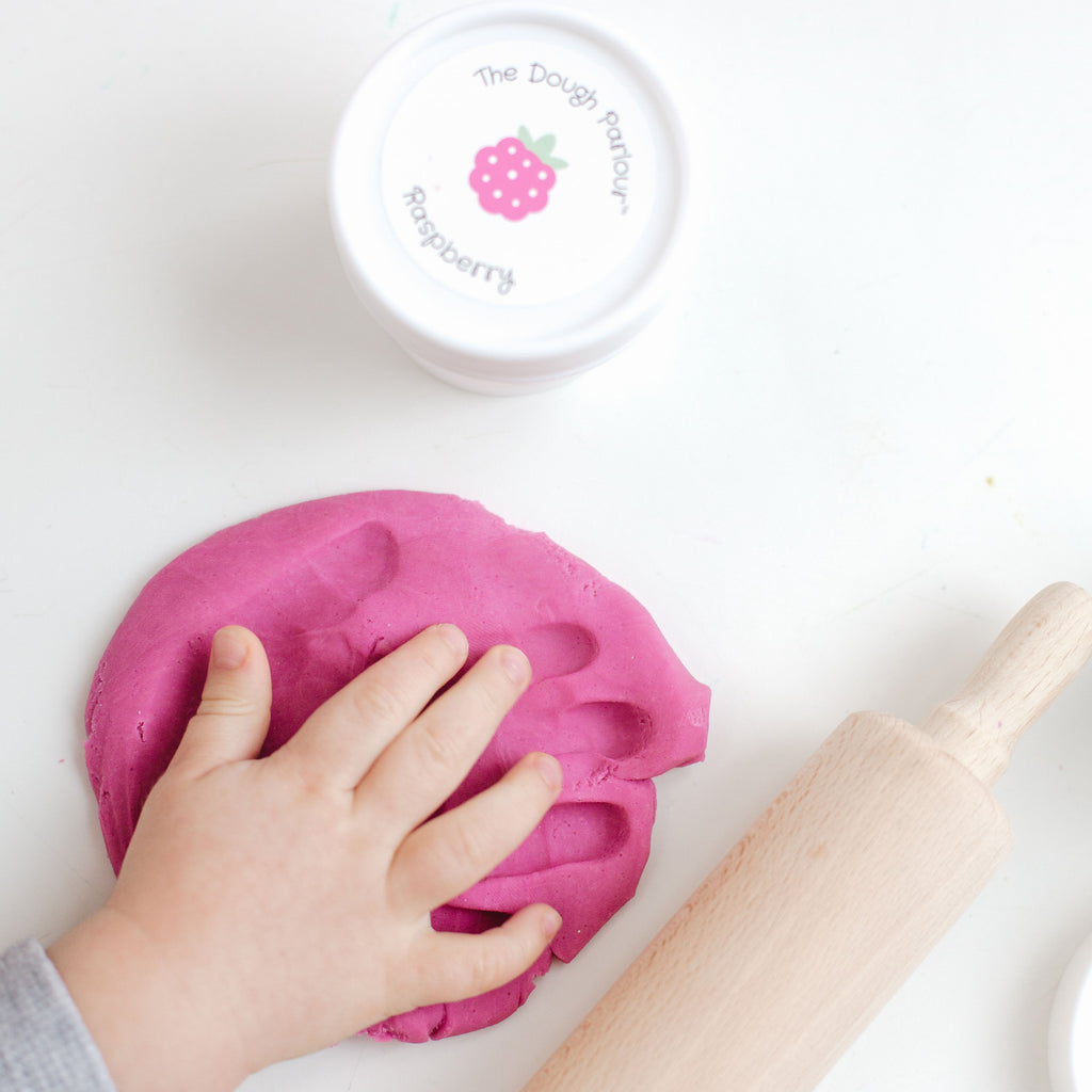 The Dough Parlour Raspberry scented pink dough tub and wooden rolling pin on white table with child's hand making a handprint in the rolled out dough